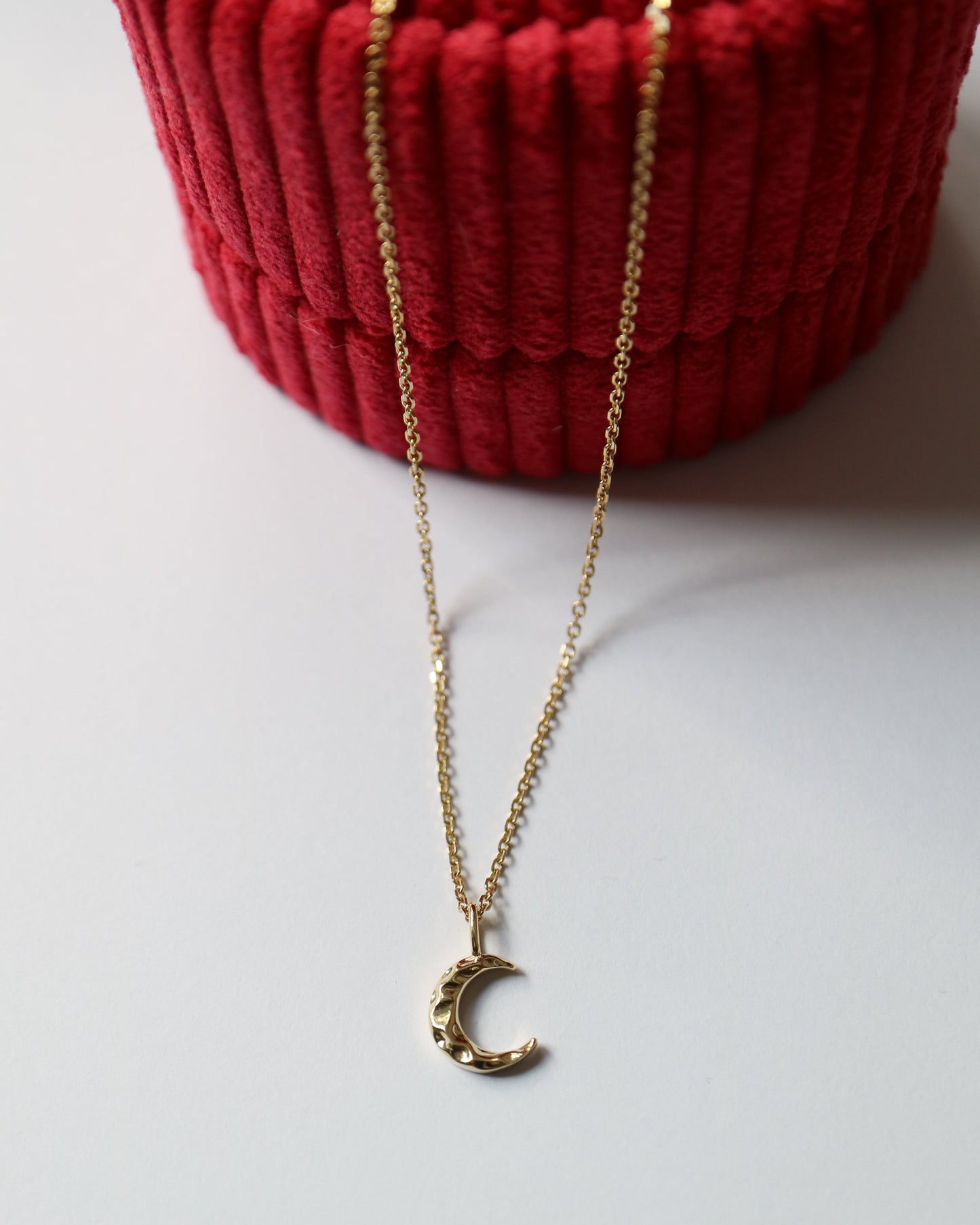 Gold necklace with a crescent moon pendant on a white surface with a red textured object in the background.