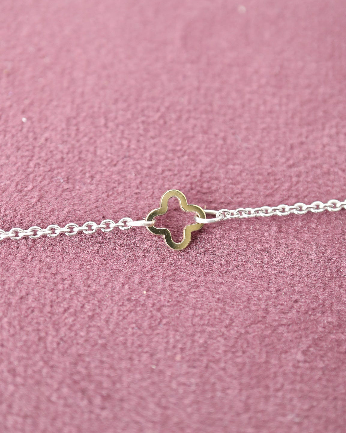 A delicate silver chain bracelet featuring a small, polished gold-toned four-leaf clover charm, displayed on a textured dusty pink velvet surface.