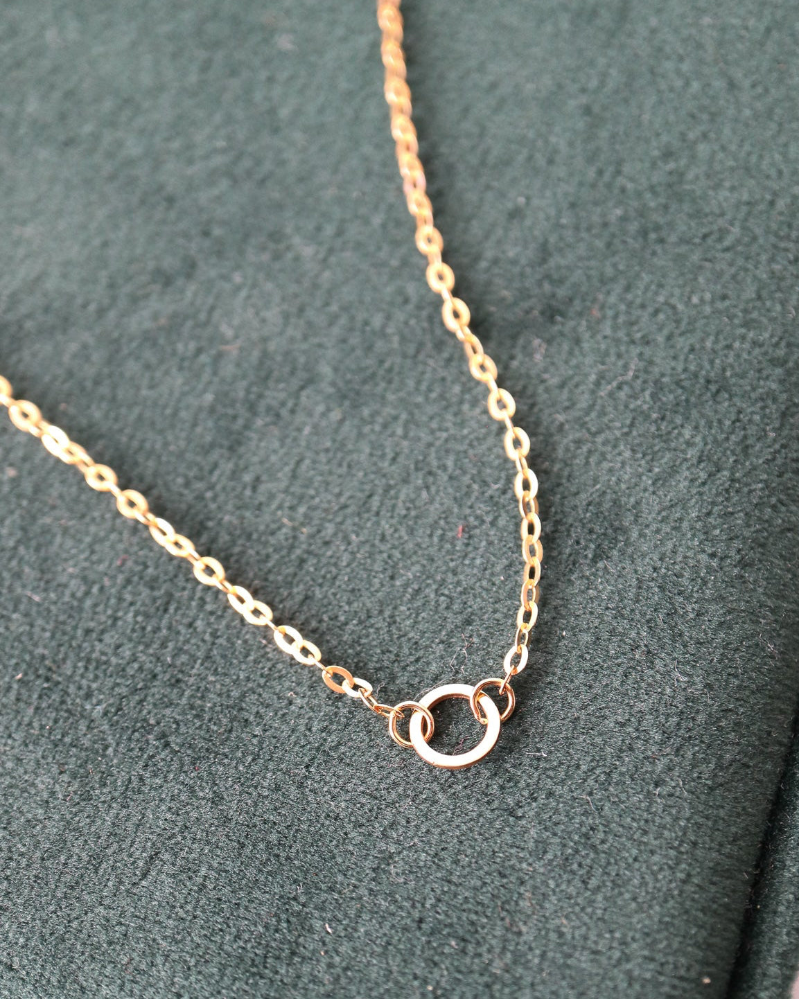 A delicate gold chain necklace with a small double-ring pendant, displayed on a dark green velvet jewelry box.