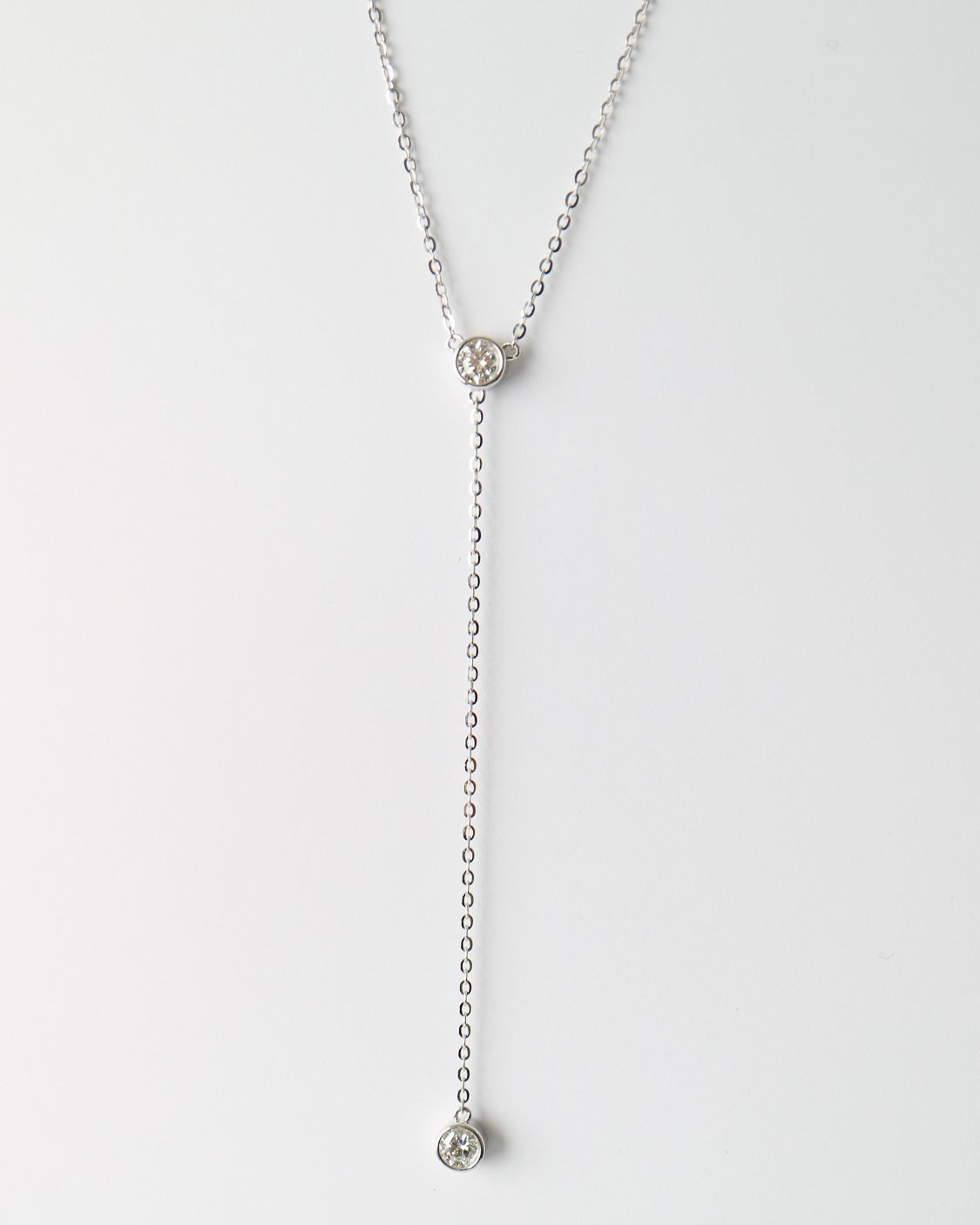 A delicate silver Y-necklace featuring a small circular floral or knot-like pendant at the top and a longer drop chain ending in a small round bezel-set stone, displayed against a clean white background.