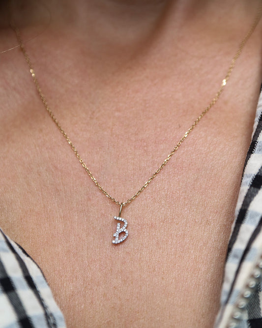 A close-up of a person wearing a delicate gold chain necklace with a small initial pendant featuring a white stone design, styled with a black and white gingham shirt.