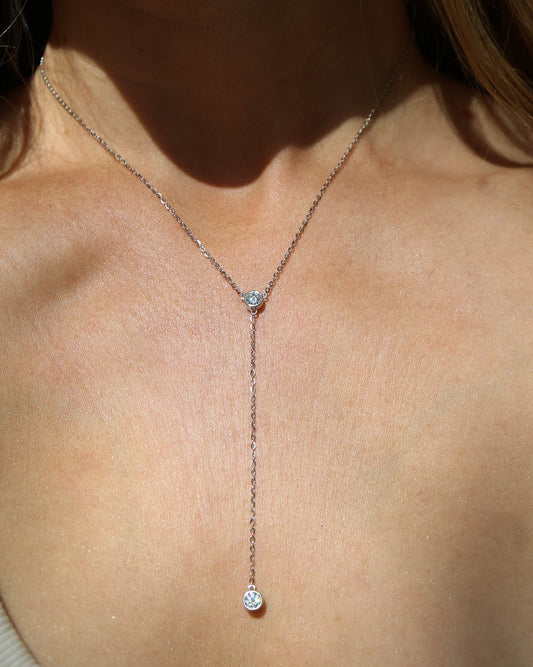 A close-up of a model's neck and chest wearing a delicate silver Y-necklace with a single small clear gemstone pendant.