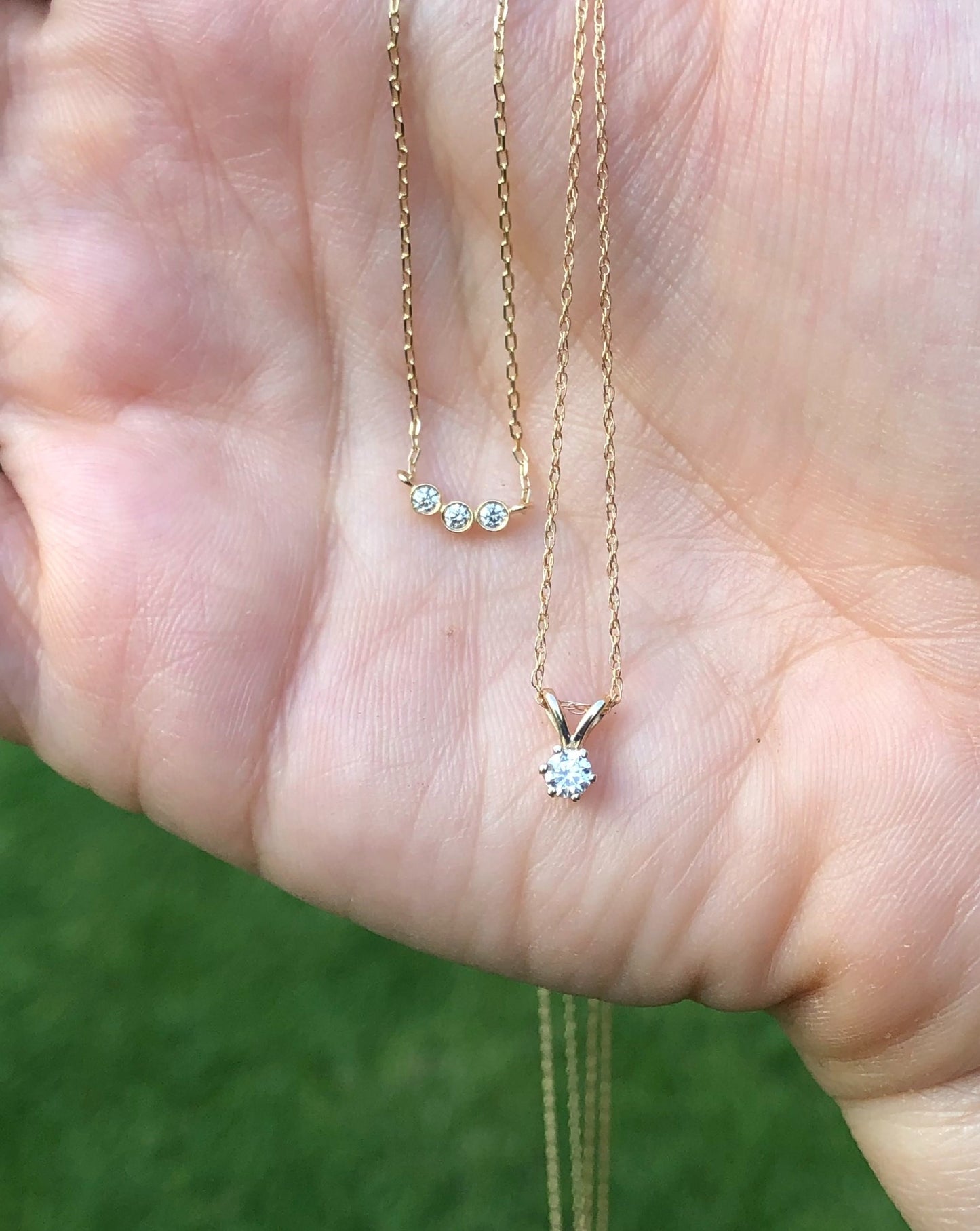 Two solid gold pendants with diamonds, shown in palm of a hand with green grass in the background