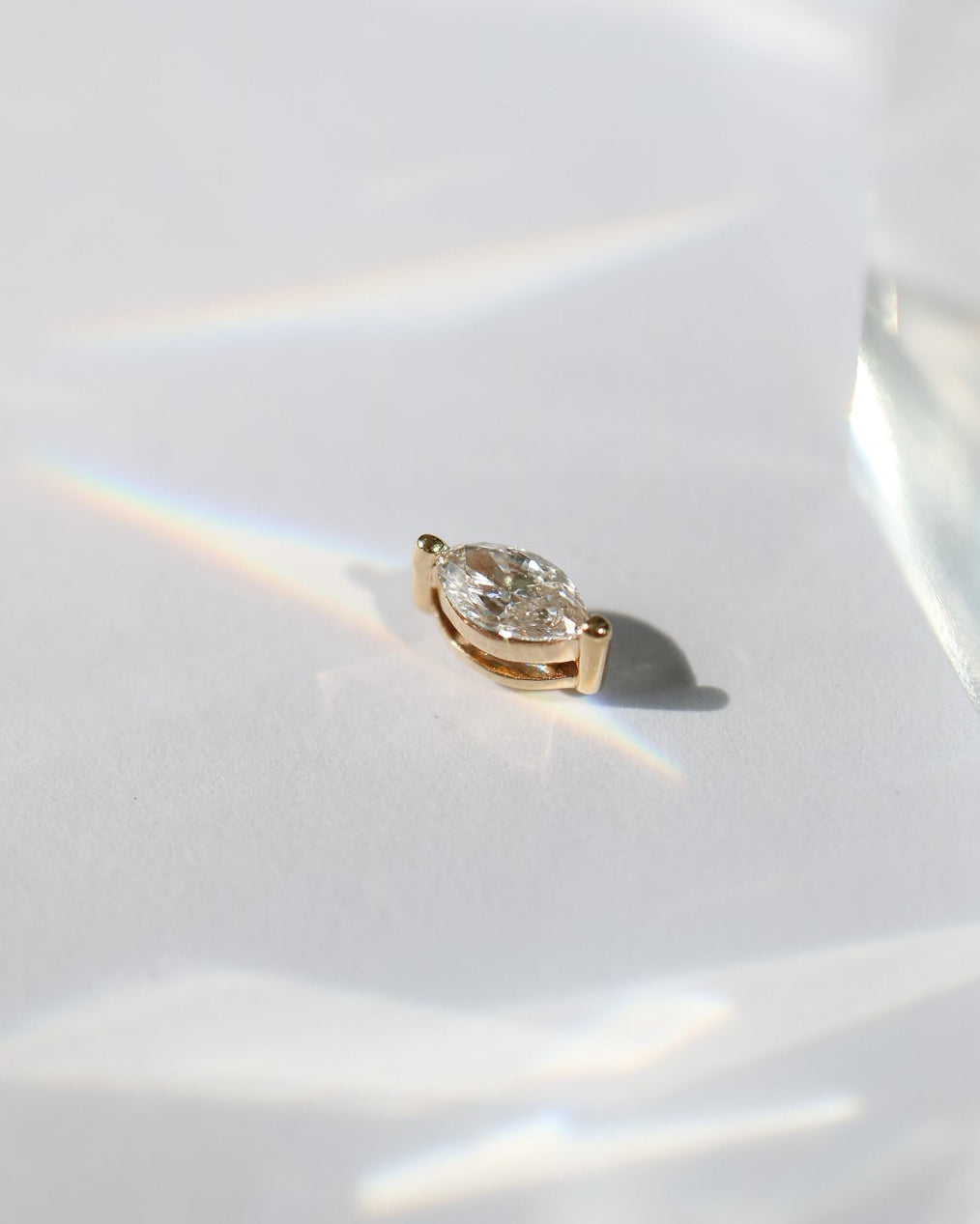 A single gold stud earring with a clear marquise diamond, photographed on a white surface with dramatic light reflections and shadows.