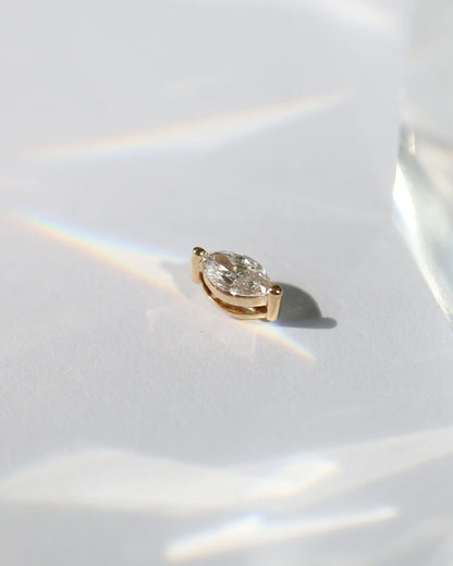 A single gold stud earring with a clear marquise diamond, photographed on a white surface with dramatic light reflections and shadows.