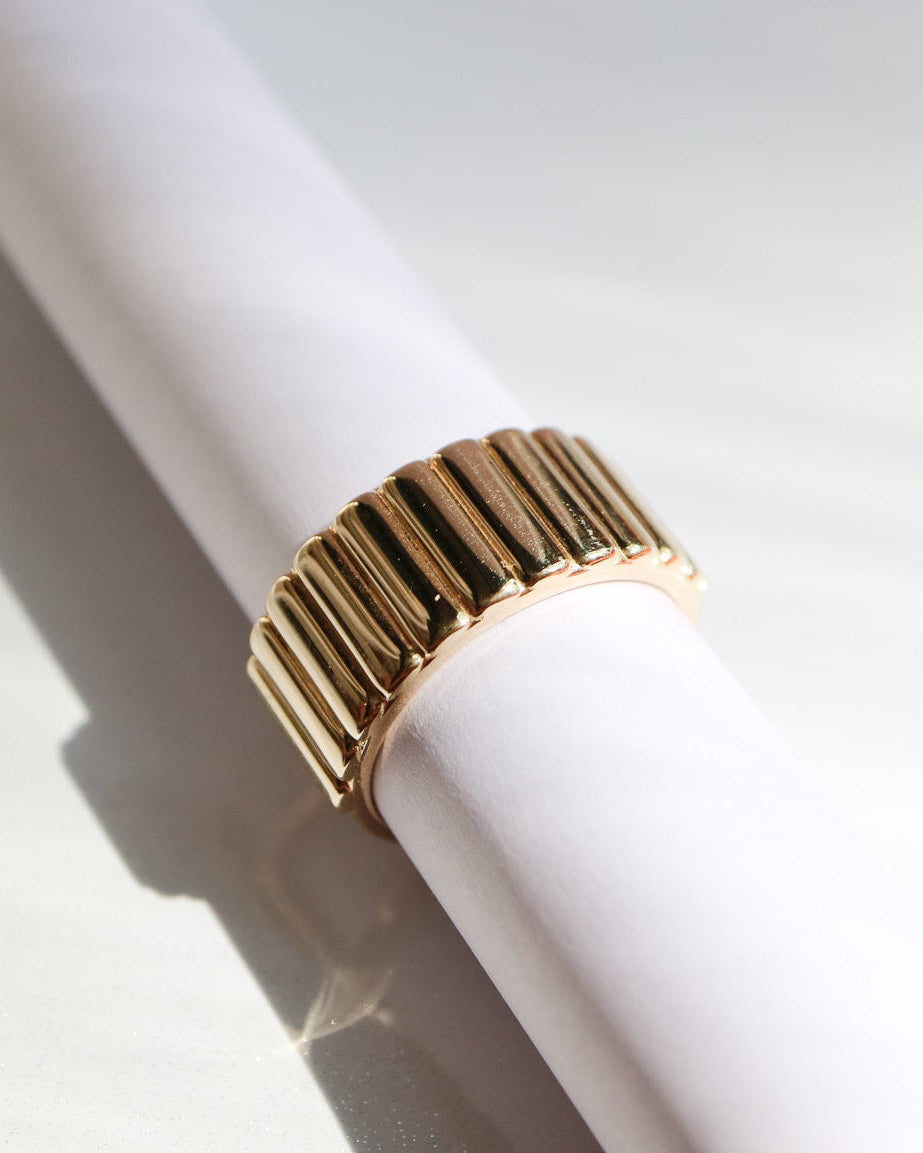 A 9ct gold segmented Oura ring jacket with a wide, flared band, displayed on a white cylindrical stand against a bright, sunlit white background.