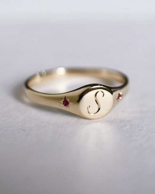 Gold ring with a central 'S' and embedded gemstones on a light gray background