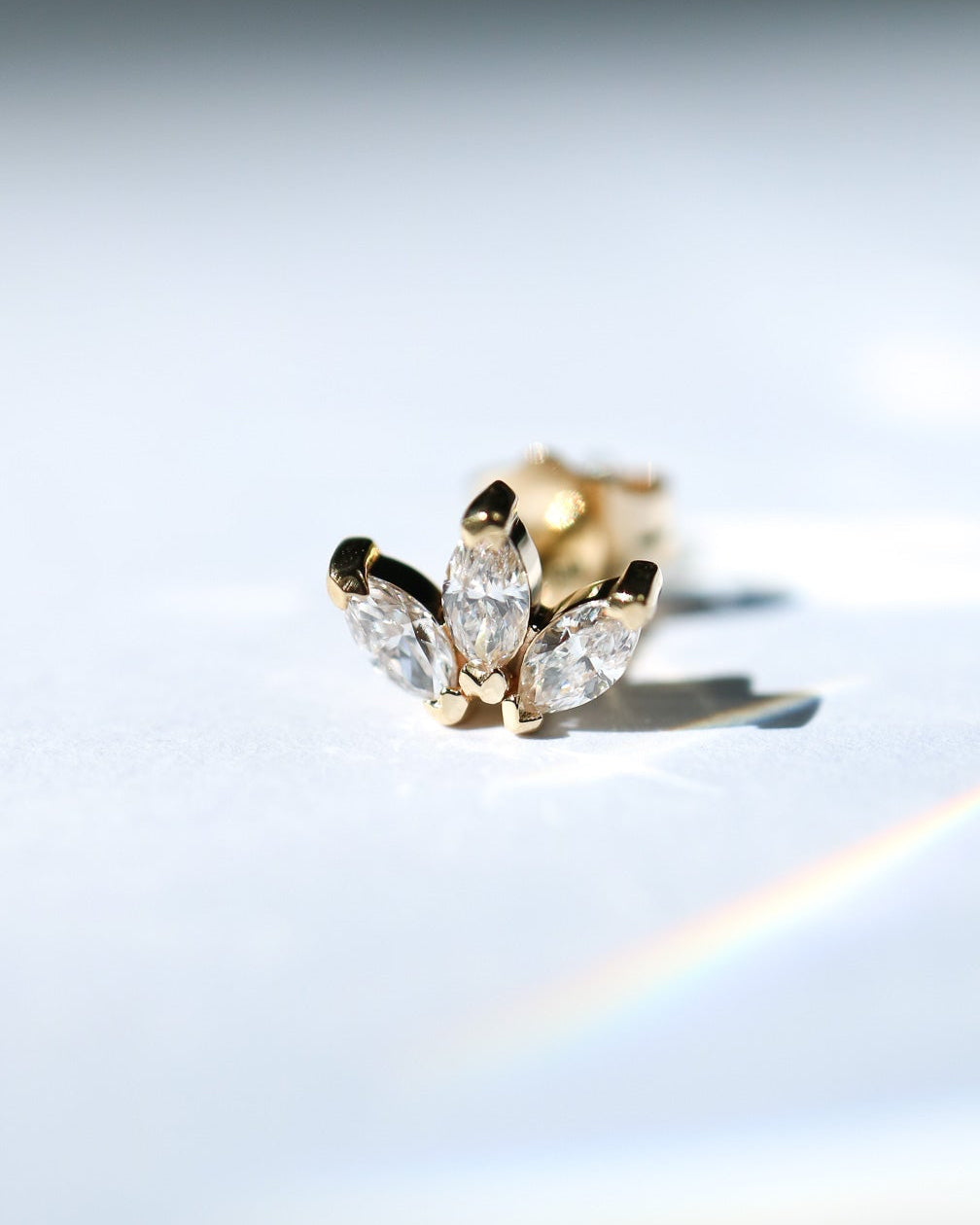 A gold stud earring featuring three marquise-cut clear gemstones in a leaf-like cluster setting, photographed on a bright white surface with soft light and a faint rainbow reflection.