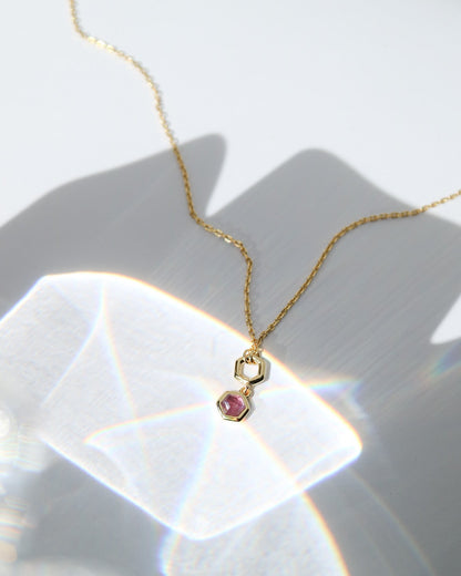 A delicate gold chain necklace with a small geometric pendant and a pink gemstone, displayed on a white surface with artistic light reflections.