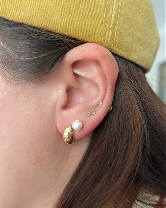 A close-up of a person's ear wearing multiple gold and pearl earrings, with a yellow cap and brown hair visible.