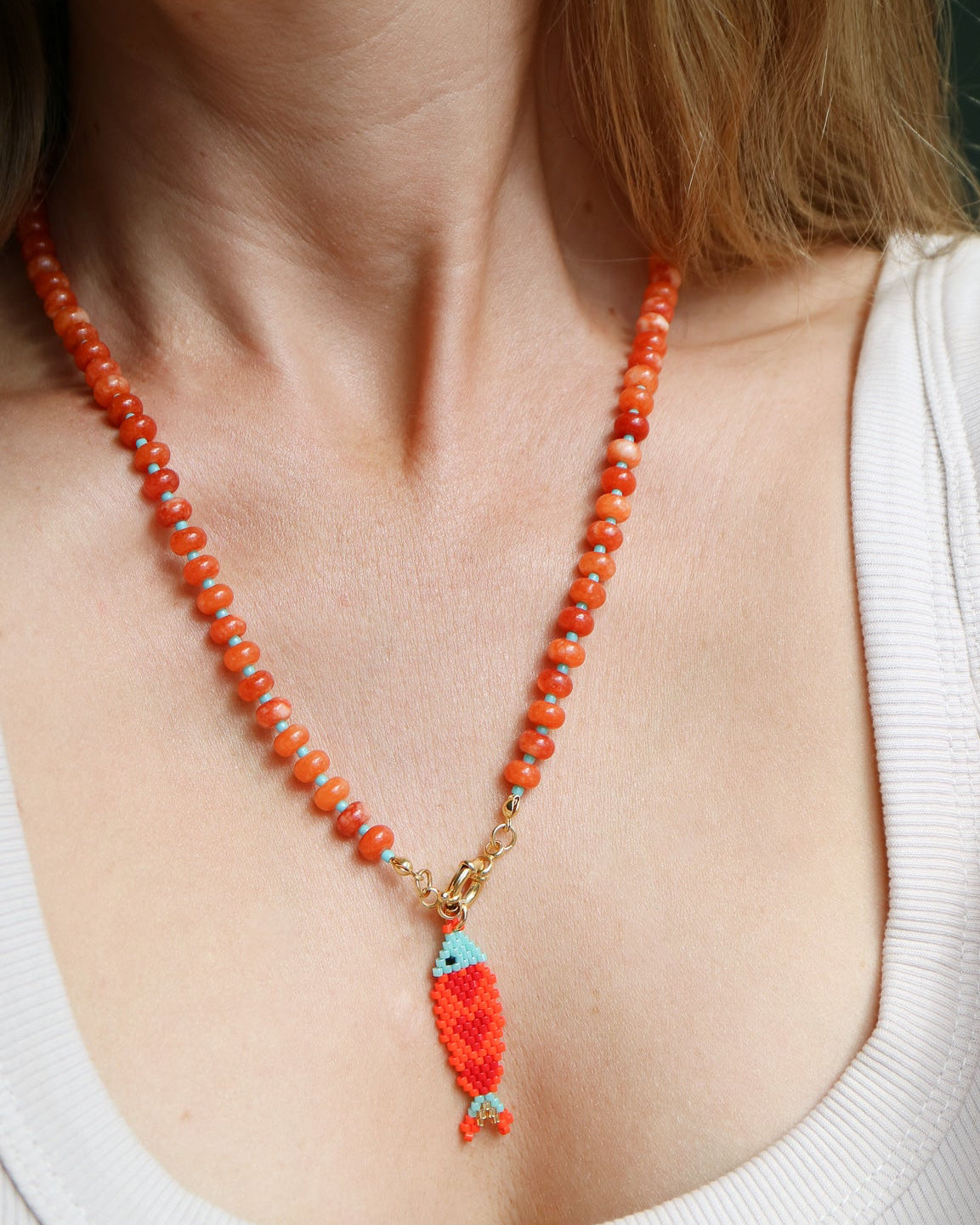 A close-up of a woman wearing a beaded necklace with orange-red beads, a gold-toned clasp, and a small fish-shaped pendant with turquoise and orange-red details.