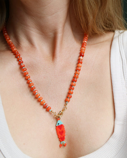 A close-up of a woman wearing a beaded necklace with orange-red beads, a gold-toned clasp, and a small fish-shaped pendant with turquoise and orange-red details.