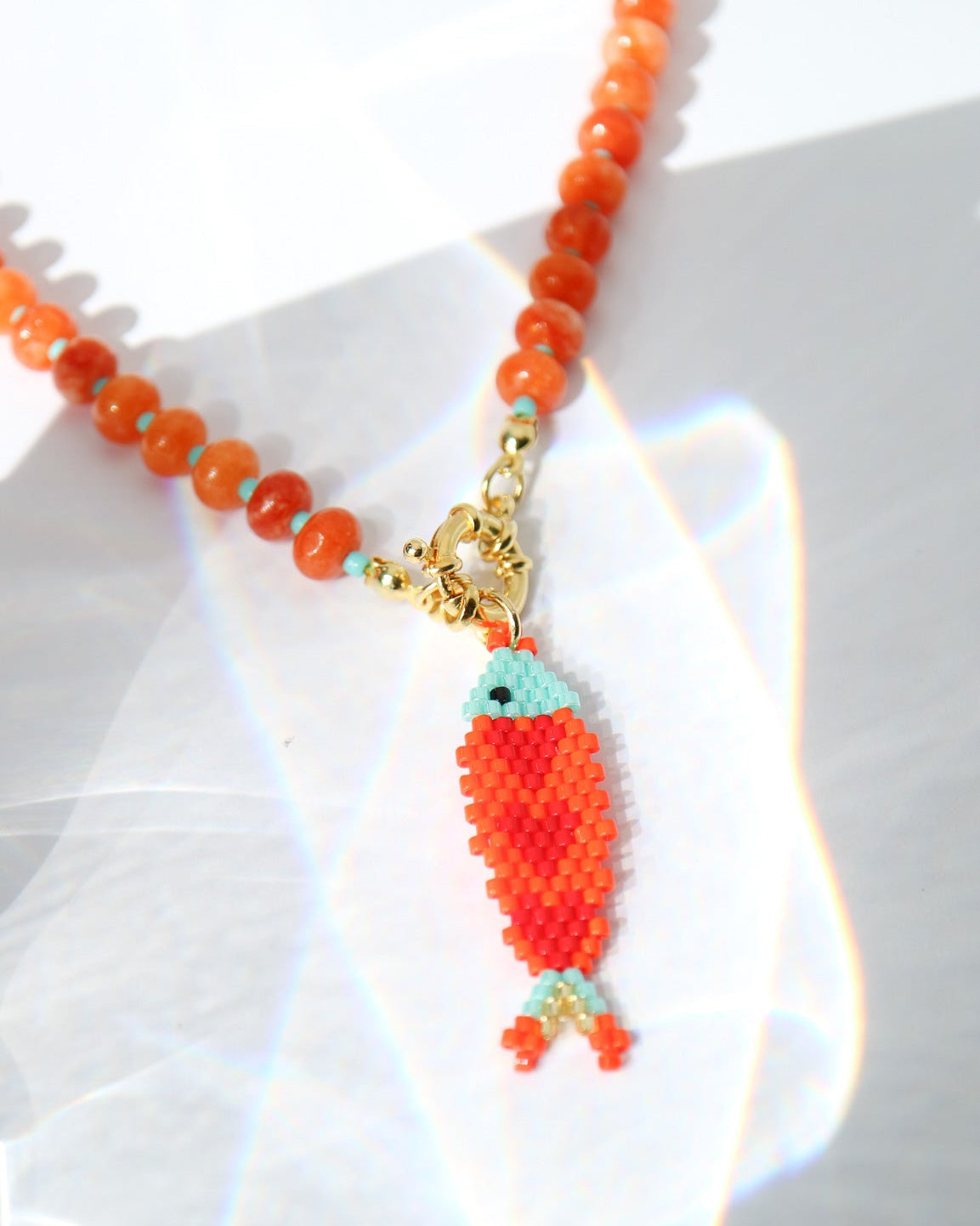 A beaded necklace with a fish-shaped pendant, featuring orange and turquoise beads with gold-toned metal accents, photographed on a white surface with sunlight and lens flare effects.