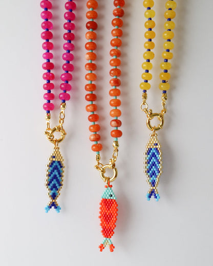 Three colorful beaded necklaces with gold-tone fish pendants, displayed vertically against a plain light background. The necklaces feature pink, orange, and yellow beads, with the center necklace's fish pendant highlighted in red-orange.