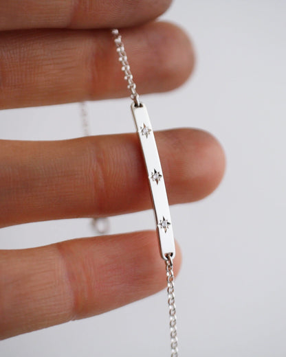 Silver bar necklace with star designs held between fingers on a light background