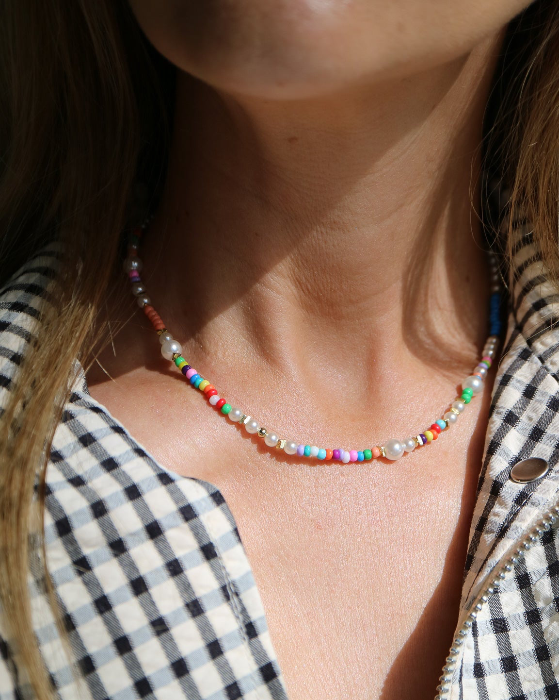 A close-up of a person wearing a short beaded necklace with colorful round beads and clear rhinestone accents, paired with a black-and-white gingham shirt.