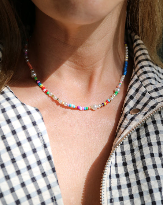 A close-up lifestyle shot of a woman wearing a delicate beaded necklace with colorful round beads and small white pearl-like accents, styled with a black and white gingham zip-up jacket.