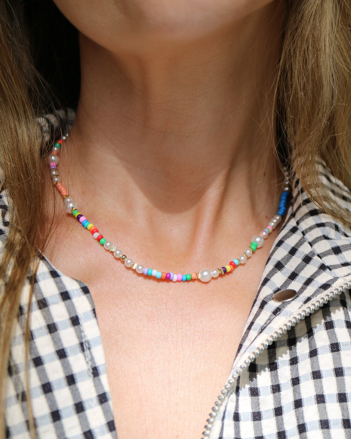A close-up of a person wearing a delicate beaded necklace with small multicolored beads and clear rhinestone accents, paired with a black and white gingham zip-up shirt.