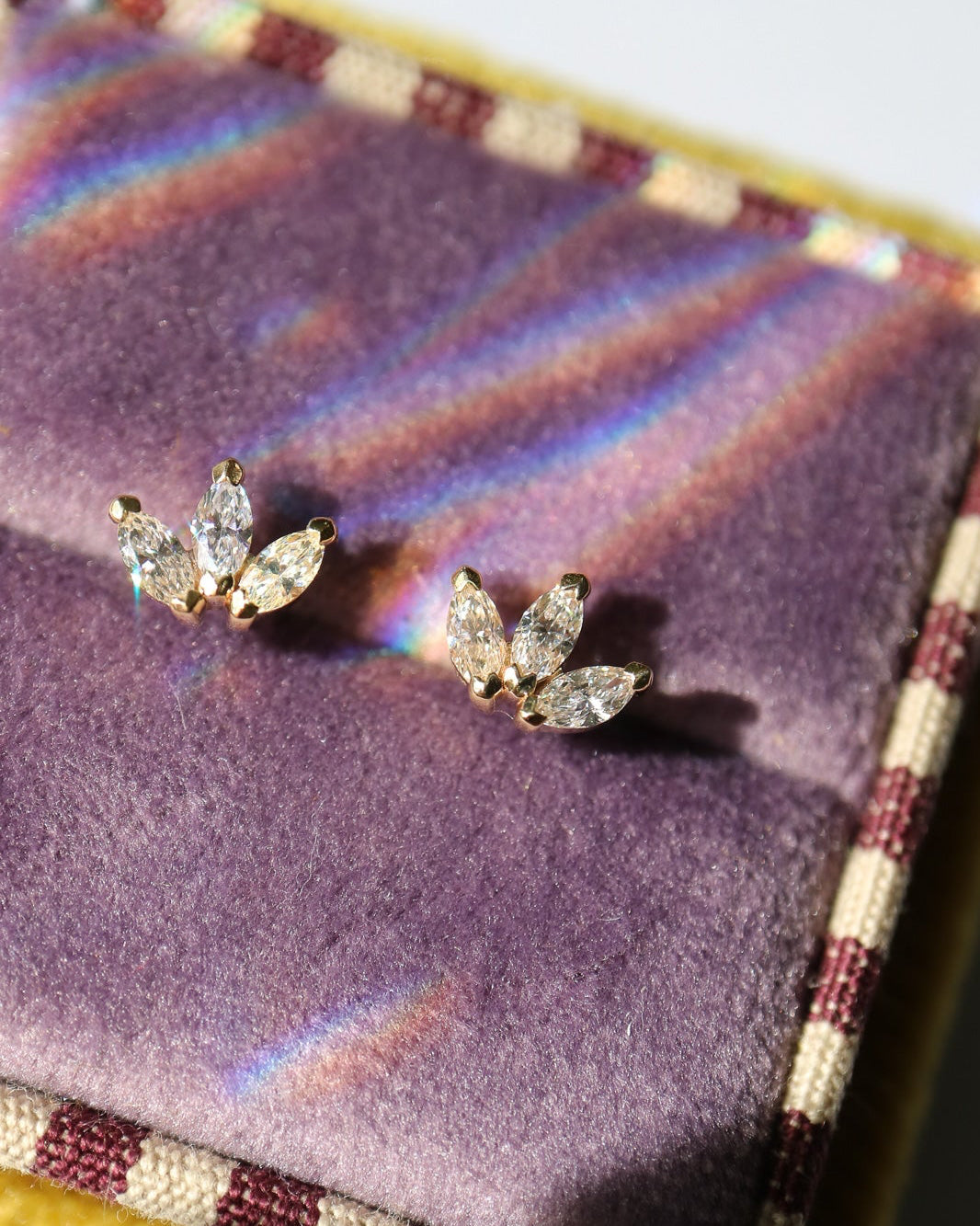 A pair of gold leaf-shaped stud earrings with marquise-cut diamonds, displayed on a purple velvet jewelry box with a patterned border.