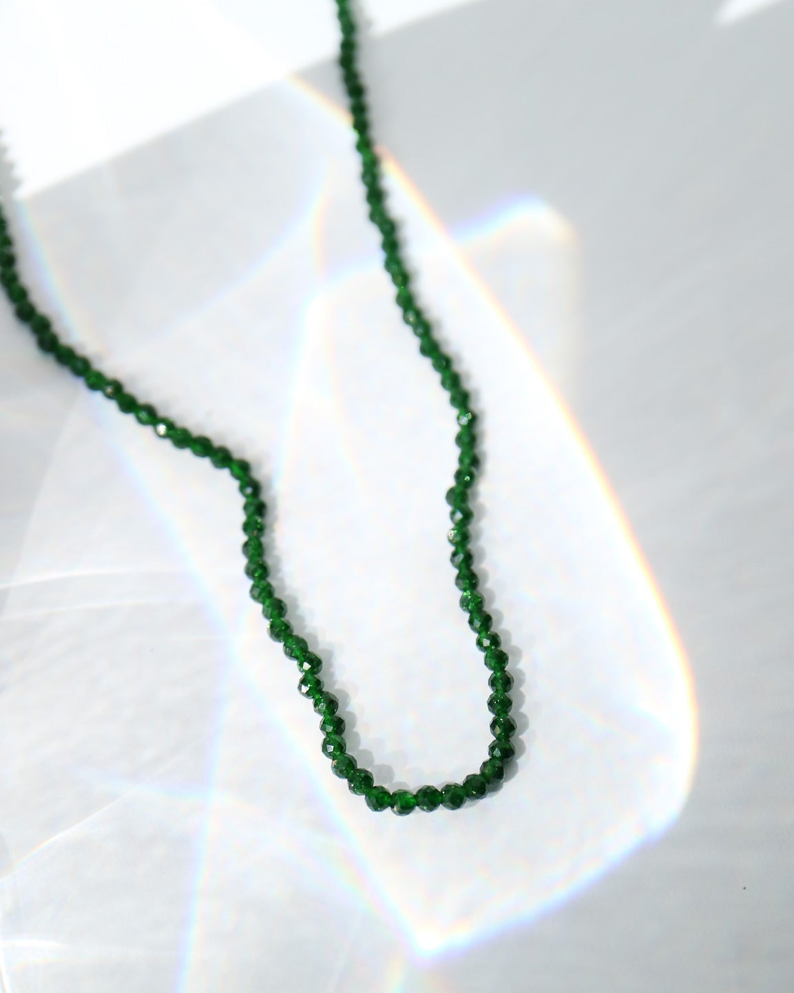 A delicate green beaded necklace draped in a loop on a glossy white surface, catching light with soft rainbow reflections.