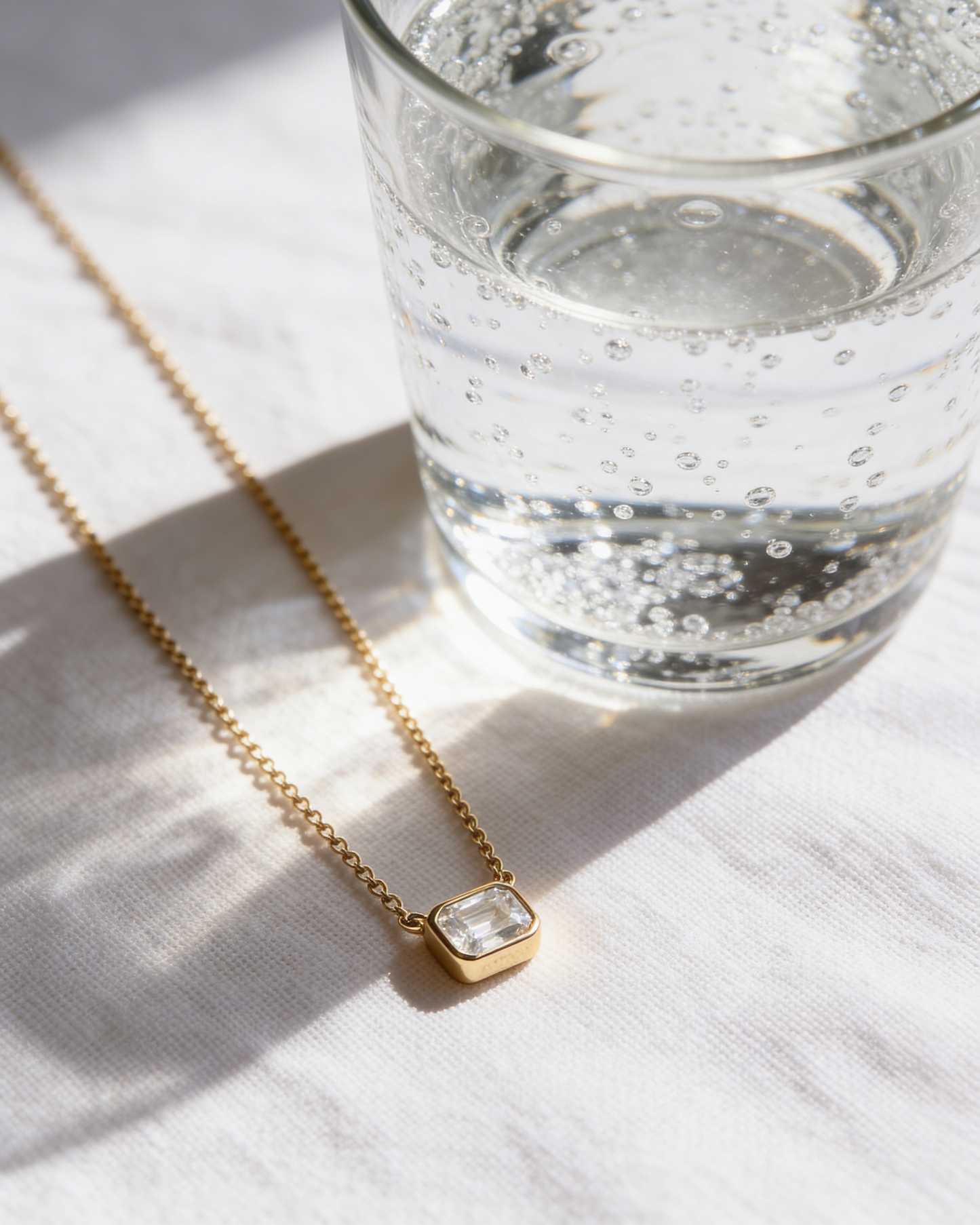 Gold necklace with a clear pendant next to a glass of water on a light surface
