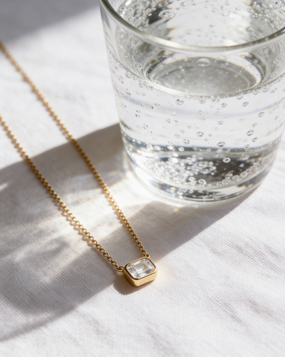 Gold necklace with a clear pendant next to a glass of water on a light surface