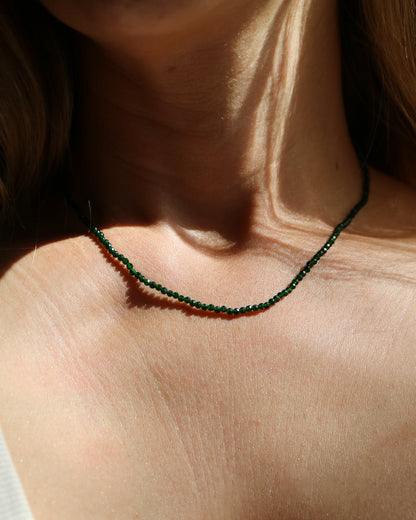 A close-up lifestyle shot of a model wearing a delicate dark green beaded necklace with small red accents, styled with a white ribbed tank top under warm, direct sunlight.