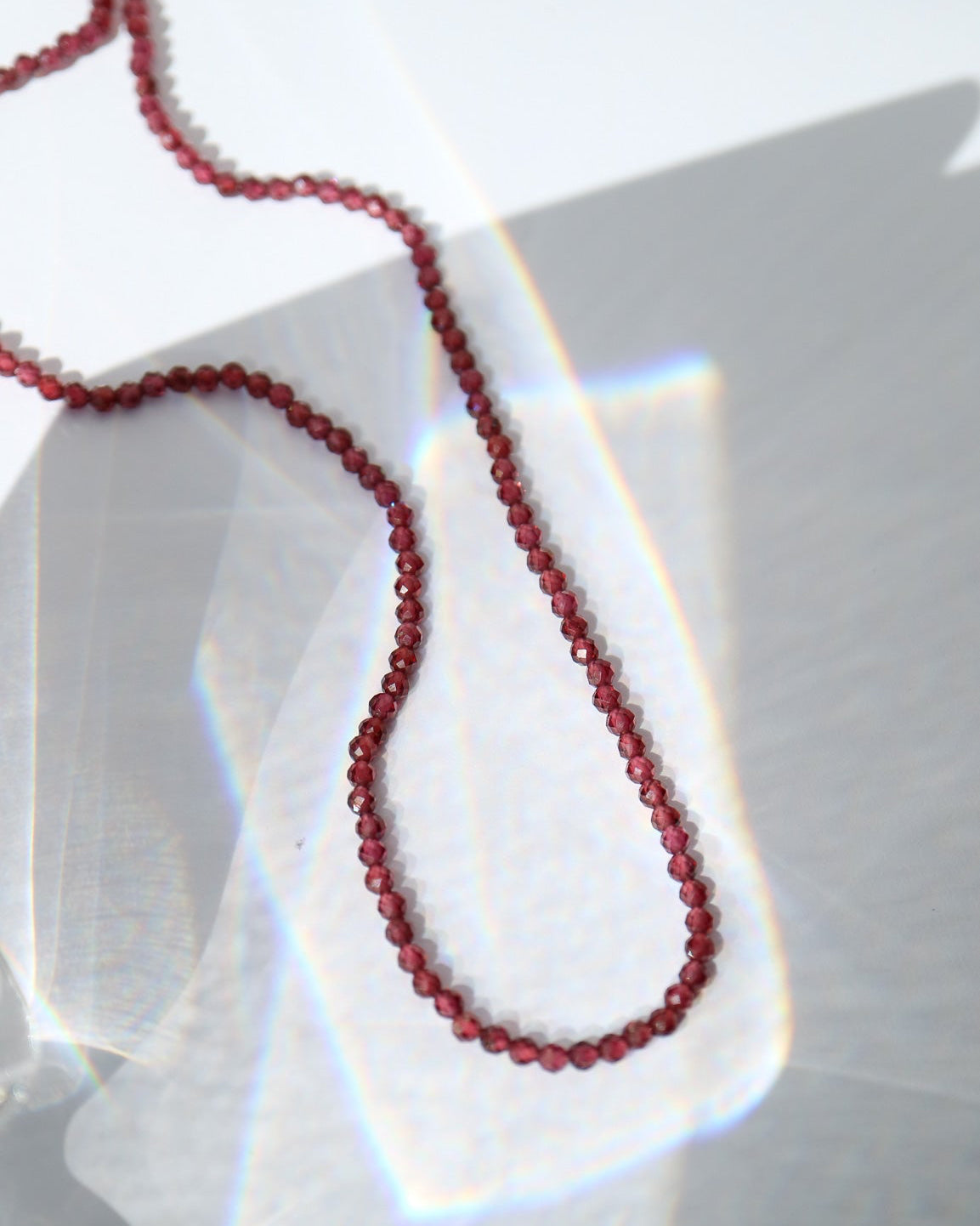 A close-up of a dark red beaded necklace draped over a translucent glass surface with dramatic light refraction and shadow patterns.