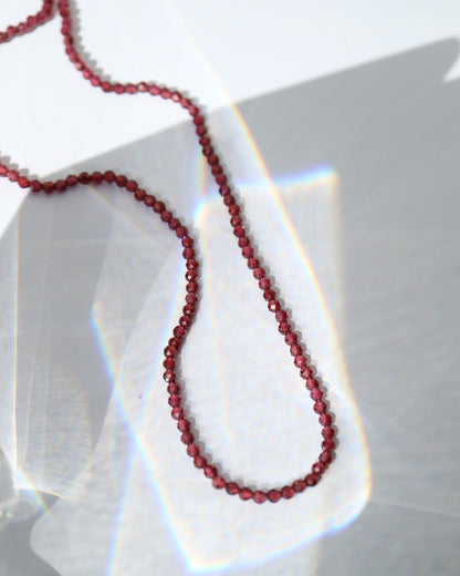 A close-up of a dark red beaded necklace draped over a translucent glass surface with dramatic light refraction and shadow patterns.