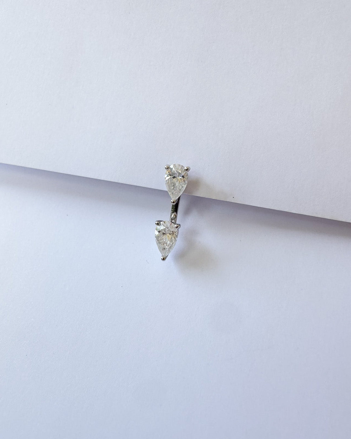 A silver-toned double drop hoop piercing ring with clear heart-shaped gemstones, resting on a plain white surface.