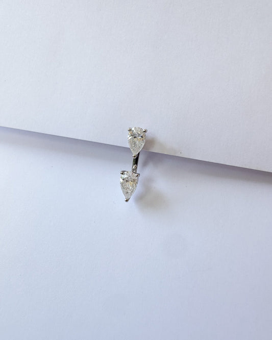 A silver-toned double drop hoop piercing ring with clear heart-shaped gemstones, resting on a plain white surface.