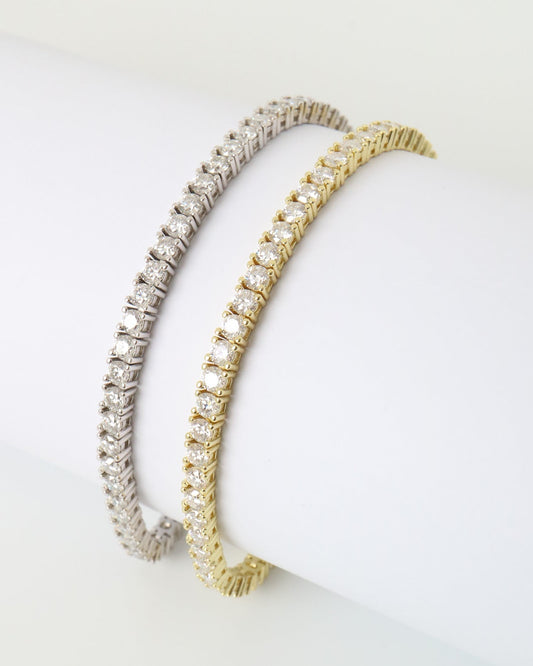 Two tennis-style bangle bracelets displayed on a white cylindrical stand, one in silver-tone metal and one in gold-tone metal, both set with clear stones.
