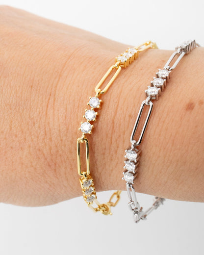 Two paperclip link bracelets on a wrist, one in yellow gold and one in silver, both featuring clear round-cut stones, shown against a plain white background.