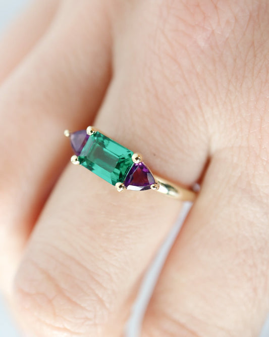 Gold ring with green and red gemstones on a finger against a light background