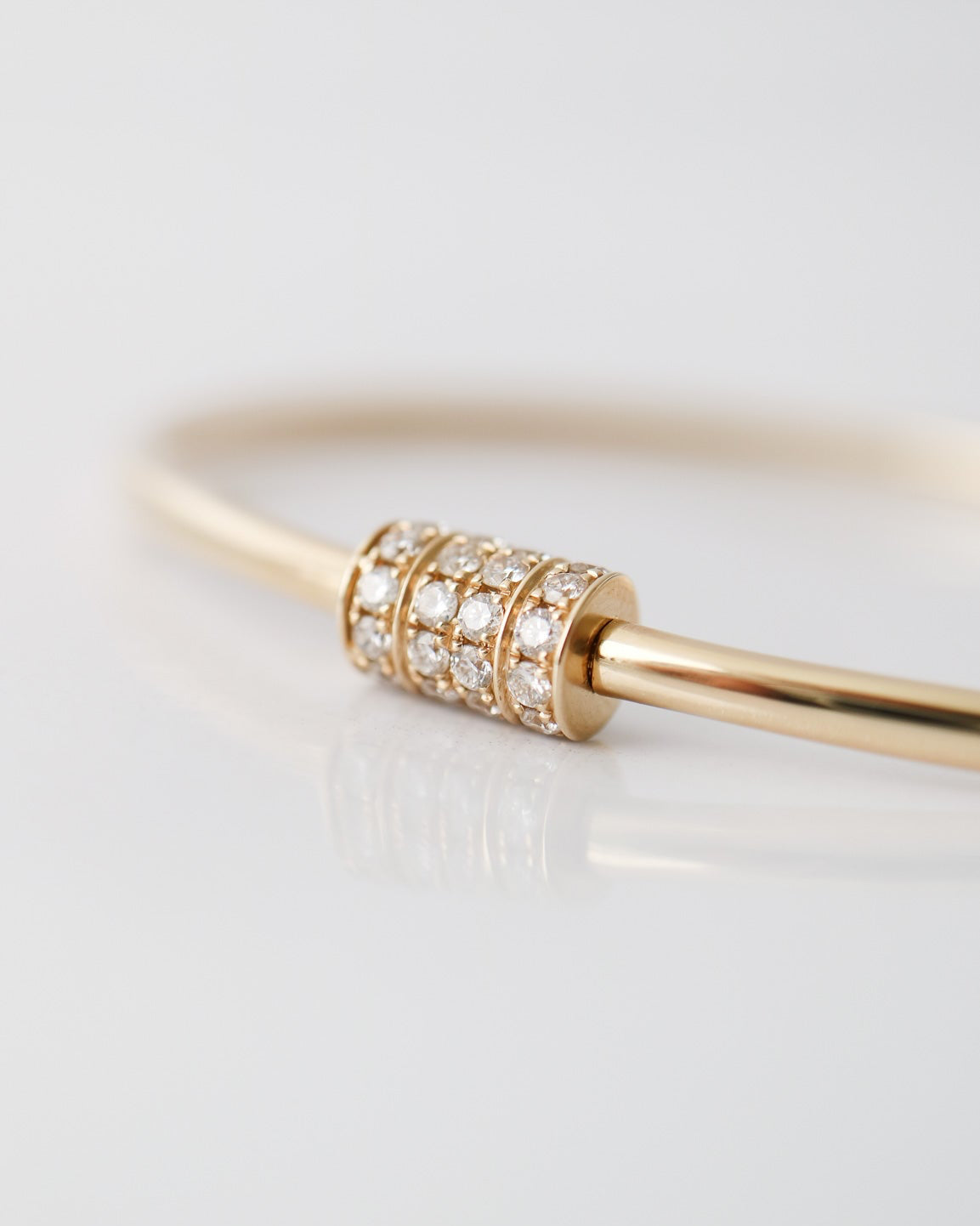 A close-up of a gold bangle bracelet featuring a central cylindrical bead set with small clear pavé stones, photographed on a clean white reflective surface.