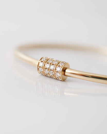 A close-up of a gold bangle bracelet featuring a central cylindrical bead set with small clear pavé stones, photographed on a clean white reflective surface.