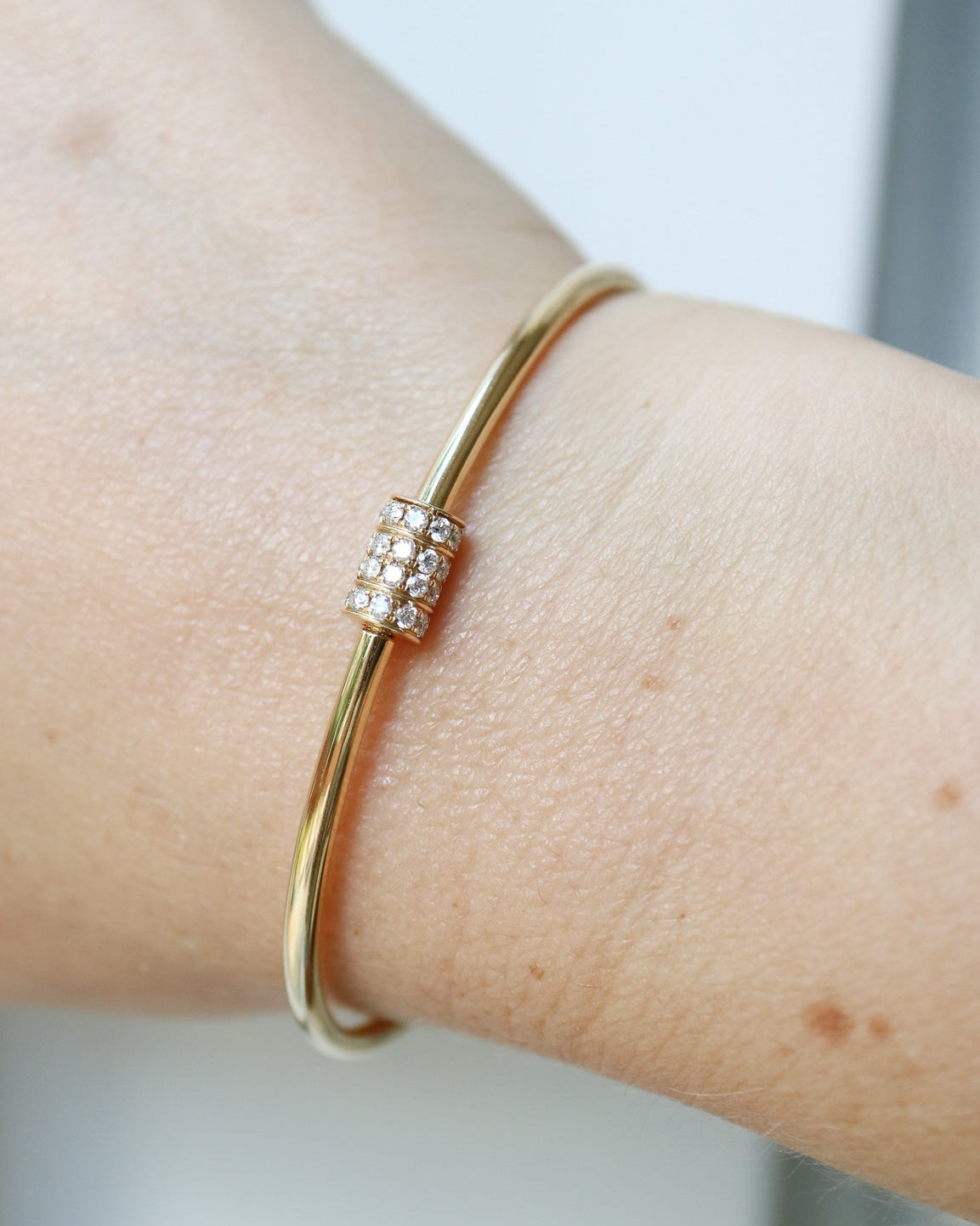 A close-up of a gold bangle bracelet with a small rectangular cluster of clear stones on the front, worn on a person's wrist.