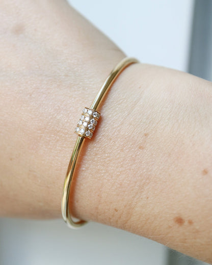 A close-up of a gold bangle bracelet with a small rectangular cluster of clear stones on the front, worn on a person's wrist.