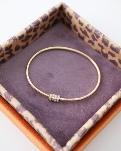 A thin gold bangle bracelet with a small pavé-set bead detail, displayed inside an open leopard-print jewelry box with a purple velvet lining on a white background.