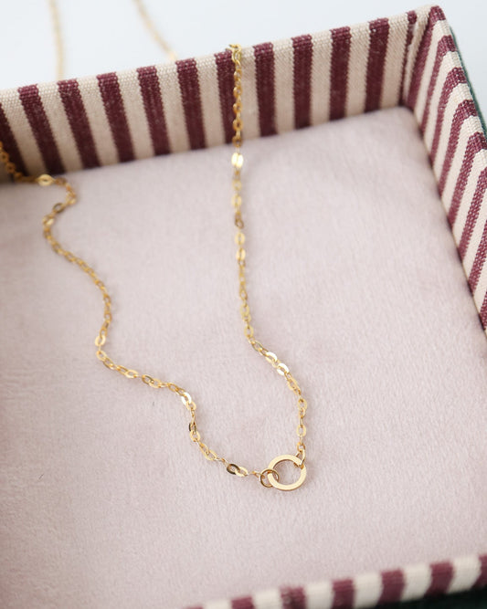 A delicate gold chain necklace with a small circular ring clasp, displayed inside a pink-lined jewelry box with a striped fabric rim.