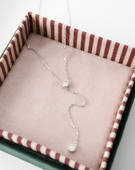 A delicate silver necklace with two small clear gemstone beads is displayed inside an open jewelry box with a pink velvet lining and a striped burgundy and cream interior.