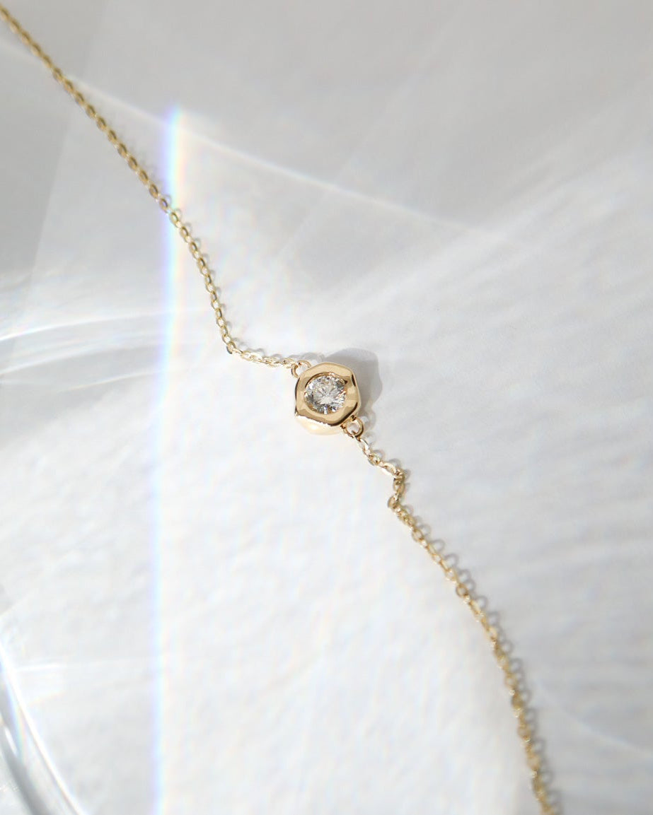 A delicate gold chain necklace with a small hexagonal gold pendant set with a clear gemstone, resting on a textured white surface with soft light reflections.