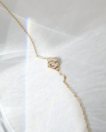 A delicate gold chain necklace with a small hexagonal gold pendant set with a clear gemstone, resting on a textured white surface with soft light reflections.