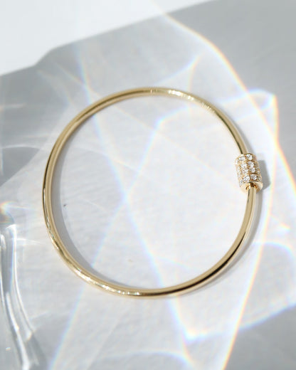 A thin gold bangle bracelet with a small pavé-set crystal accent on the clasp, displayed on a white surface with soft light leaks.