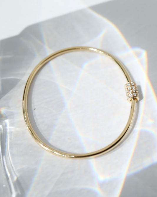 A thin gold bangle bracelet with a small pavé-set crystal accent on the clasp, displayed on a white surface with soft light leaks.
