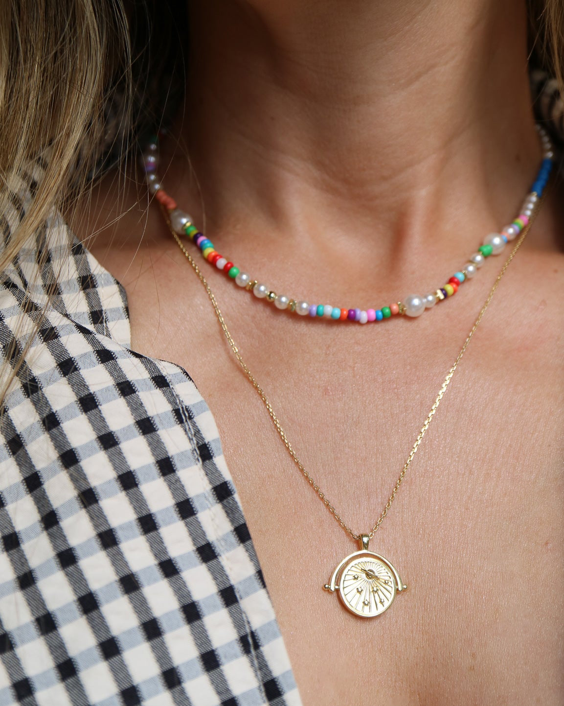 Close-up of a person wearing layered necklaces: a short beaded choker with multicolored beads and white pearls, and a delicate gold chain necklace with a round clock-face pendant.