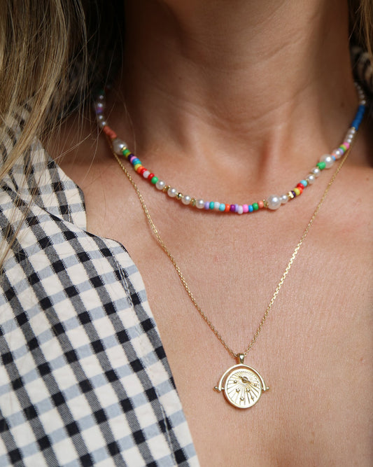 Close-up of a person wearing layered necklaces: a short beaded choker with multicolored beads and white pearls, and a delicate gold chain necklace with a round clock-face pendant.