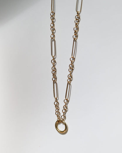 A gold-toned chain necklace featuring elongated oval links and a small circular ring pendant, displayed against a plain light gray background.