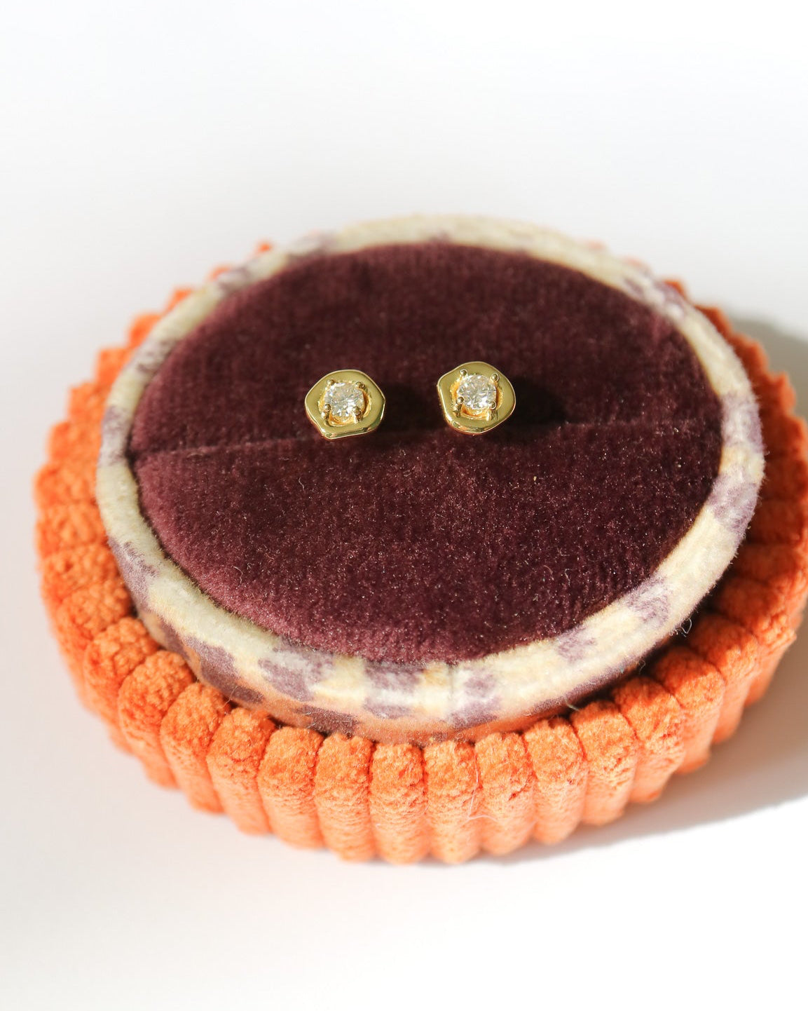 A pair of small gold stud earrings with clear stones, displayed on a round burgundy velvet jewelry stand inside an orange ribbed holder against a white background.