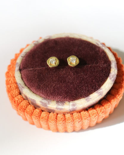 A pair of small gold stud earrings with clear stones, displayed on a round burgundy velvet jewelry stand inside an orange ribbed holder against a white background.